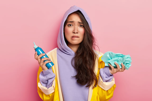 Image of frustrated young woman bites lower lip, holds spray and handkerchief, looks nervously at camera, feels sick, caught cold during rainy day, wears purple hoody. Autumn, people, health problem