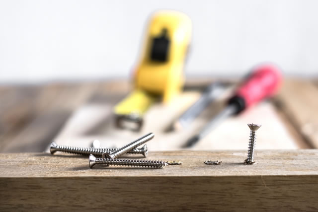 set of silver screws on wooden top table background. One of them screwed into timber wood and blurred screw driver, measuring tab and pencil in the back