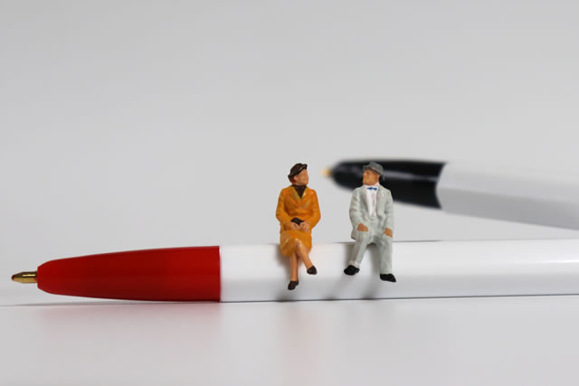 A miniature old couple sitting on a red ballpoint pen with a black ball pen