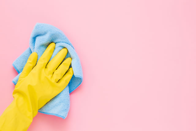 Employee hand in yellow rubber protective glove wiping pastel pink wall from dust with blue dry rag. General or regular cleanup. Commercial cleaning company. Copy space. Empty place for text or logo.