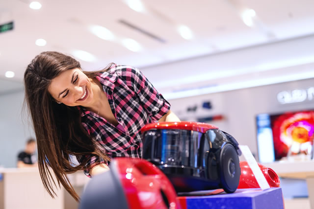 Smiling gorgeous Caucasian brunette with long hair dressed casual looking for new vacuum cleaner while standing in tech store.