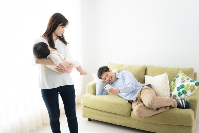 赤ちゃんを抱っこしながらソファでスマホをしている夫にあきれているような女性